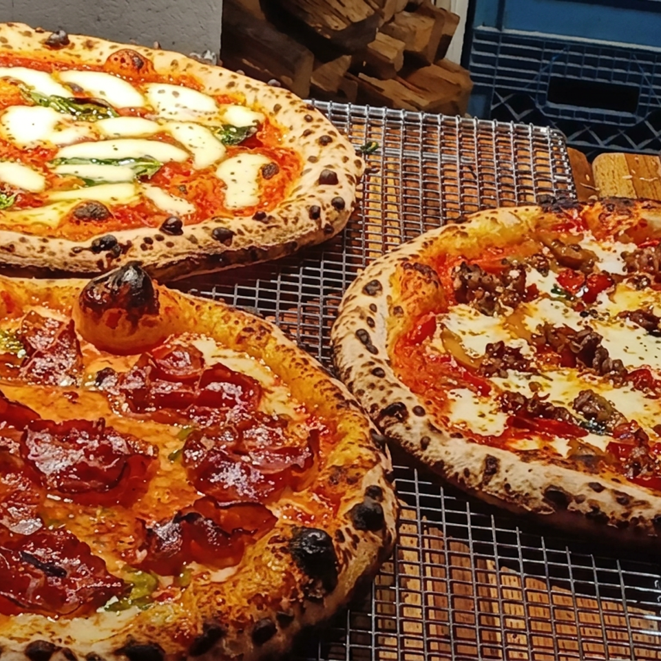 Three wood-fired pizzas with blistered crusts, melted cheese, tomatoes, basil, pepperoni, and ground meat cooling on wire racks.