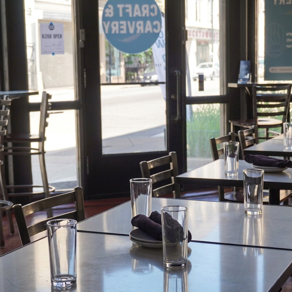 Sunlit restaurant dining room with neatly set tables, water glasses, black napkins, and large street-facing windows.